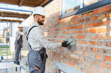 İşçi dış tuğla duvara malayla sıva uygularken diğer bir işçi arka planda inşaat işi yapıyor.