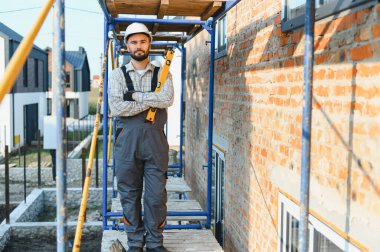 İnşaat işçisi, inşaat başlığı ve tulumlarıyla iskelede duruyor, bir inşaat sahasında ruh seviyesini tutuyor.