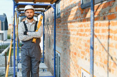 Kasketli ve tulumlu bir adam inşaat sahasında ruh seviyesinde bir alet tutuyor, tuğla bir duvar inşa ediyor.