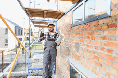 İnşaat işçisi, kasketli ve tulumlu, havan topu tutan, tuğla duvarın yanında duran.