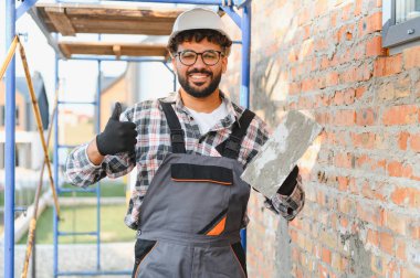 Genç duvarcı, kasketli ve tulumlu, havan topu tutan, tuğla duvarın yanında duran, başarıyı inşa eden