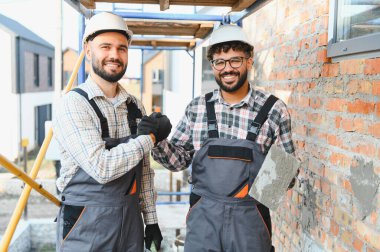 Sert şapka ve tulum giyen inşaatçılar, elinde malayla yeni ev inşaatında başarılı bir takım çalışmasını kutluyorlar.