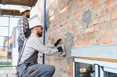 İnşaat işçileri tuğla duvara sıva uyguluyor. Ekip binanın ön cephesinde çalışıyor. El emeği ve becerilerini gösteriyor.