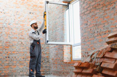İnşaatçı yeni pencere kuruyor. İnşaat halindeki tuğladan bir duvardaki ruhsal seviyeyle mükemmel bir dikey hizalama sağlıyor.