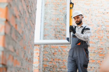 İnşaat işçisi sabit bir şapka ve tulum giyerek tam olarak yeni bir pencere çerçevesini ruh seviyesini kullanarak tuğla bir duvara yerleştiriyor.