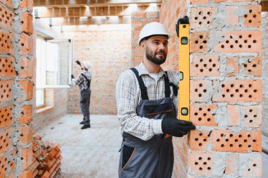 İnşaatçı sabit şapka ve tulum giyerek inşaat sahasında duvar düzlüğünü sağlıyor. Pencere kuran başka bir işçi