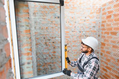 İnşaat işçisi, yenilenme sırasında tam hizalama için ruh seviyesini kullanarak tuğla duvar açıklığına pencere çerçevesi kuruyor.