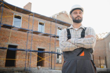 Sert şapkalı ve tulumlu sakallı adam çapraz kollu, konut inşaatını denetliyor.