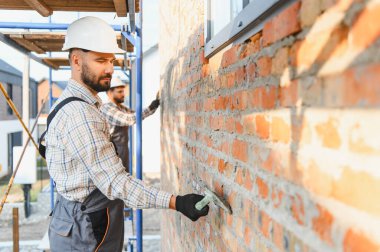 İnşaat işçileri iskelede duvara havan topu uyguluyor, bir ev inşa ediyorlar. Kasketli adamlar şantiyede çalışıyor.