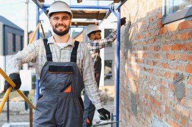 Profesyonel inşaat işçisi gülümsüyor, sert bir şapka takıyor ve inşaat iskelesinde tulum giyiyor, bir meslektaşı da tuğla duvara sıva yapıştırıyor.