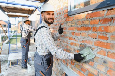 Mason işçisi tuğla duvar örüyor, havan topunu malayla sürüyor, gülümsüyor ve iş memnuniyeti için el sallıyor.