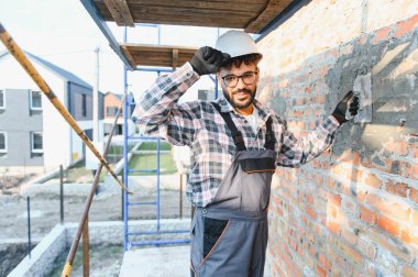 Kasketli ve tulumlu genç adam inşaat alanına tuğla örgü örüyor, kameraya gülümsüyor.