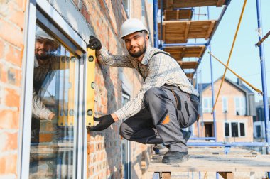 İnşaat işçisi tuğla duvardaki ruh seviyesini kullanarak pencere çerçevesini kontrol ediyor, bina kurulumu sırasında hassasiyet sağlıyor