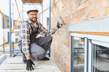 İnşaat işçisi kasketli, kırmızı tuğlalı dış duvarı iskeleye yapıştırıyor, gülümsüyor ve inşaat çalışmalarına odaklanıyor.
