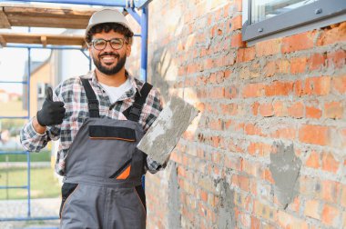 Sert şapkalı ve tulumlu bir adam havan topuyla bir mala sarılmış, baş parmak işareti yapıyor, başarıyı ve iyi işi simgeliyor.