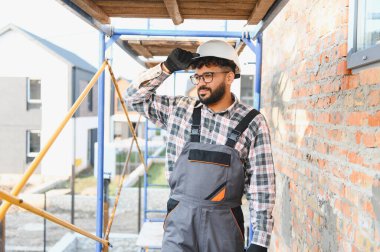 İnşaat işçisi yeni konut inşaatı projesini denetliyor. Güvenlik kıyafeti, sıkı bir şapka ve tulum giyiyor.