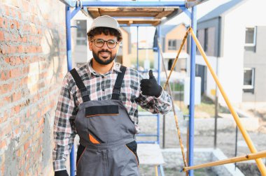 Gülümseyen inşaat işçisi, sert şapka ve tulum giyiyor, başparmak işareti yapıyor, başarıyı ve memnuniyeti gösteriyor.