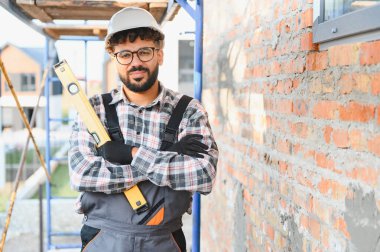Kasketli, ekose gömlekli ve tulumlu genç bir müteahhit. Kolları katlanmış, tuğla duvara yaslanmış.
