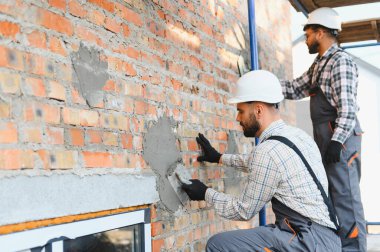 İki yetenekli inşaat işçisi kırmızı tuğla duvara sıva uyguluyor, tadilat ve inşaat işleri yapıyorlar.