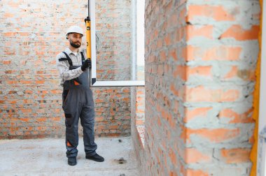 Sert şapkalı ve tulumlu bir adam inşaat alanındaki tuğladan bir duvarın içine yeni bir pencere çerçevesi yerleştiriyor.