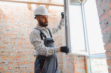 Sert şapkalı ve tulumlu genç adam tuğla bir duvara yeni bir pencere yerleştiriyor, tadilat yapıyor.