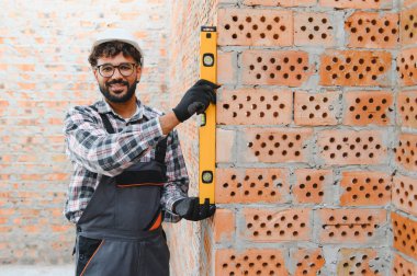 Gülümseyen inşaat işçisi, kasketli ve tulumlu, ruhsal seviyedeki tuğla duvarın hizasını kontrol ediyor.