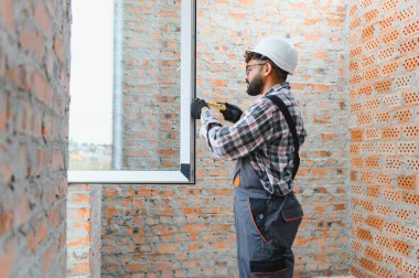 İnşaat işçisi bir inşaat alanındaki tamamlanmamış tuğla bir duvara yeni bir pencere çerçevesi yerleştiriyor ve ölçüyor. Ev geliştirme kavramı