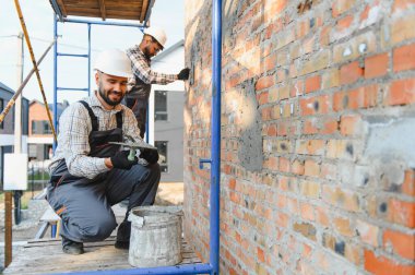 İki inşaat işçisi yeni bir ev duvarını sıva ve tuğlayla örüyor, kalın şapkalar takıyor ve iskele üzerinde çalışıyor.