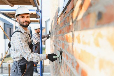İnşaat işçisi, sert şapka ve güvenlik eldiveni takıp inşaat alanındaki bir malayla tuğla duvara havan topu atıyor.