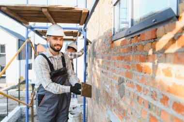 Güvenlik miğferi takmış inşaatçı yeni bir binanın dış duvarını sıva ve iskeleyle kaplıyor.