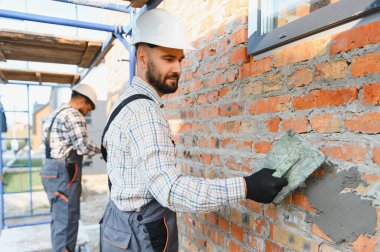 Sert şapkalı ve tulumlu masonlar yeni bir ev duvarına havan topu sürüyor. İnşaatı, takım çalışmasını ve ev yapımını sembolize ediyor.