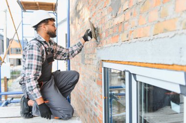 Sert şapka takan ve tulum giyen sakallı bir adam iskeledeki dış tuğla duvara alçı yapıştırıyor.