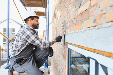 İnşaat işçisi, baret ve güvenlik gözlüğü takıyor, inşaat sahasında malası olan tuğla bir duvara titizlikle havan topu atıyor.