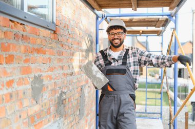 İnşaat işçisi kasketli ve tulumlu, betonla bir mala tutan, tuğla duvarın ve iskelenin yanında duran mutlu.