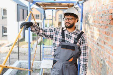 Kasketli ve tulumlu genç bir müteahhit inşaat sahasında iskeleye çıkıp tuğla bir duvar inşa ediyor.