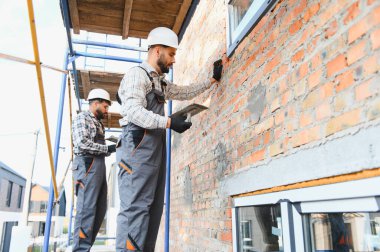 İnşaat işçileri iskeledeki tuğladan duvara sıva uyguluyor, bir bina yenileme projesi üzerinde çalışıyorlar.