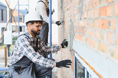 İki inşaat işçisi iskele üzerinde çalışırken mala kullanarak dış tuğla bir duvara alçı uyguluyor.