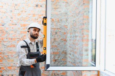 Sert başlıklı inşaat işçisi, pencere iskeleti seviyesini tuğla duvara karşı kontrol ederek, kesin ev kurulumunu sağlama alıyor