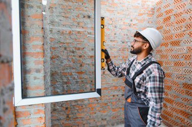 Sert şapka ve tulum giyen yetenekli işçiler, pencere çerçevesinin tuğla duvarda düzgün hizalandığından emin olmak için ruh seviyesini kullanıyorlar.