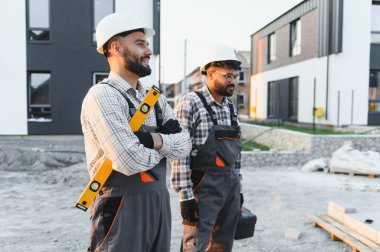 İnşaat alanında duran, takım çalışmalarını ve profesyonel inşaat çalışmalarını gösteren sert şapka ve tulum giyen işçiler
