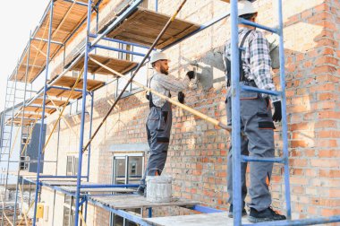 İnşaat işçileri eski bir tuğla duvara sıva uyguluyor. İskeleden, evin dışını yeniliyorlar.