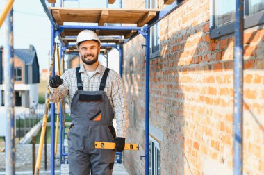 Kasketli ve tulumlu gülümseyen inşaat işçisi inşaat sahasında ruhsal düzeyde bir alet tutuyor.