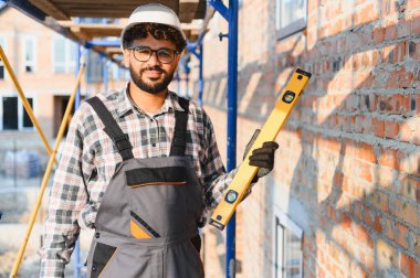 Başlık ve tulum giyen, ruh seviyesini yüksek tutan, inşaat alanında tuğla bir duvarın önünde duran bir mimar.