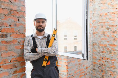 Kendine güveni tam bir müteahhit, iş tulumu giyiyor, tamamlanmamış tuğla duvarlı bir odada duruyor.