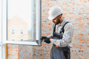 İnşaat işçisi, inşaat başlığı ve tulumuyla tuğla duvar açılışında tornavida kullanarak pvc pencere çerçevesi kuruyor.