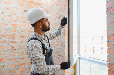 Kasketli ve üniformalı inşaat işçisi modern plastik bir pencereyi tuğla bir duvardaki bir deliğe yerleştiriyor. Ev yenileme konsepti