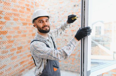 Gülümseyen inşaat işçisi tuğla bir duvara pencere yerleştiriyor, koruyucu eldiven ve şapka takıyor.