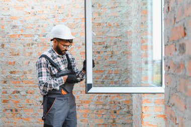 Sert şapkalı ve iş elbiseli bir adam, tornavida kullanarak tuğla duvar açılışına modern bir pencere çerçevesi kuruyor.