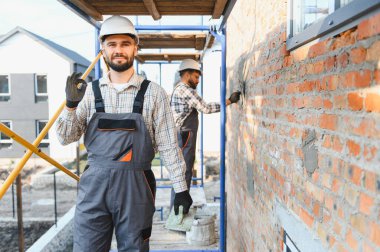 Başlık ve tulumlar içinde inşaat işçisi, başka bir inşaatçının tuğla duvarı sıvadığı bir tabela yapıyor.