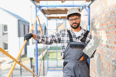 Kubbe ve güvenlik donanımı takan usta inşaat işçisi iskeleye çıkıp tuğla bir duvar inşa ediyor. El emeği konsepti